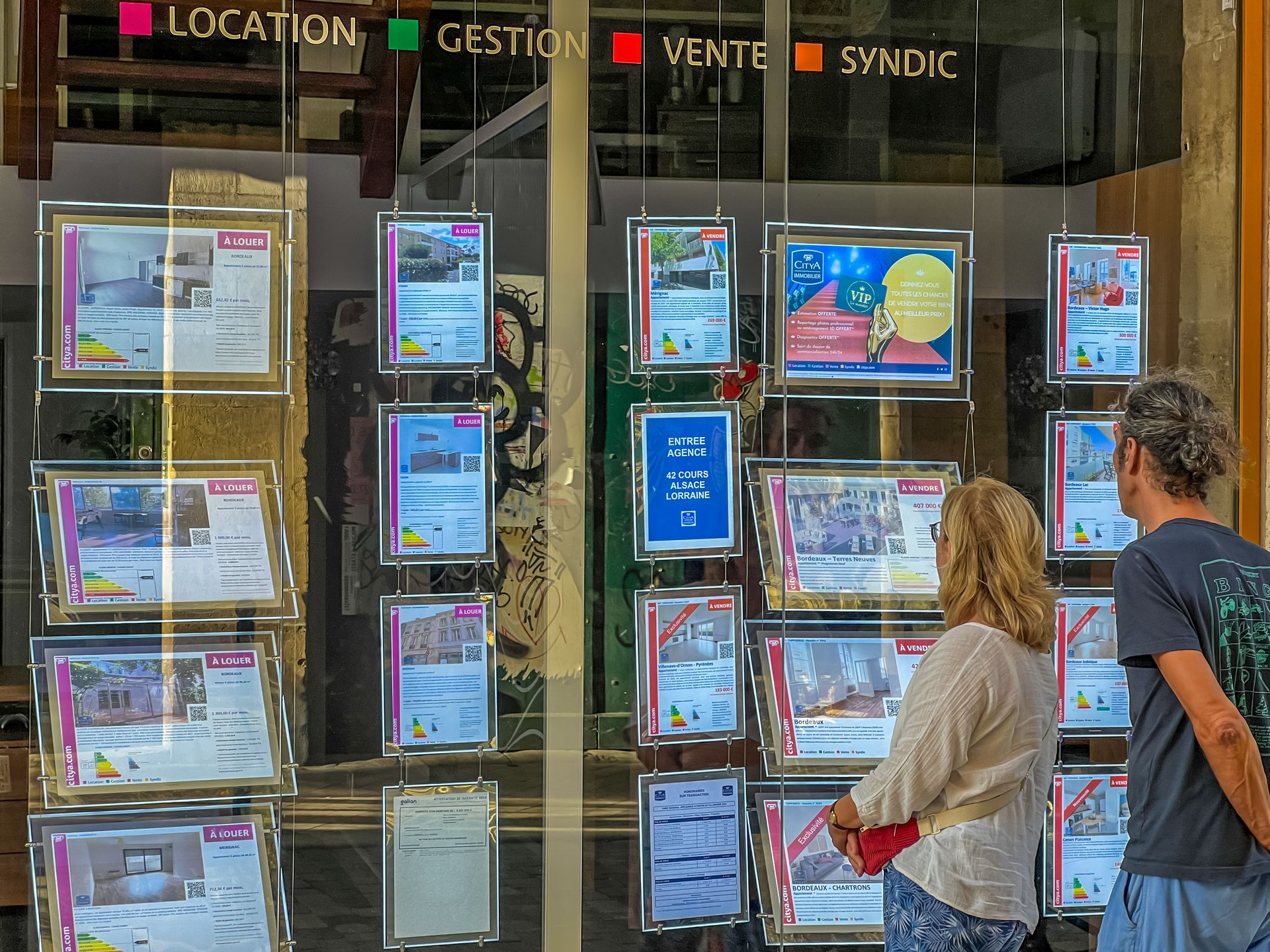 Un couple consulte des annonces immobilières dans vitrine d’agent immobilier, Bordeaux.